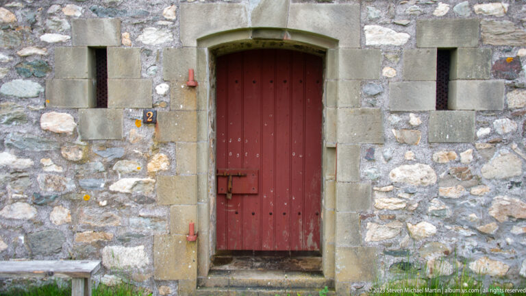 Red plank door with iron hinges and latch in a dressed-stone surround, flanked by narrow openings in a fieldstone wall.
