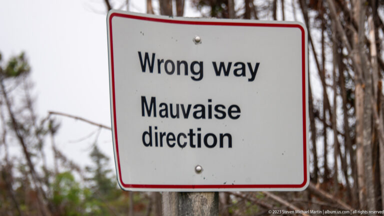White bilingual sign reading “Wrong way / Mauvaise direction” beside woodland.