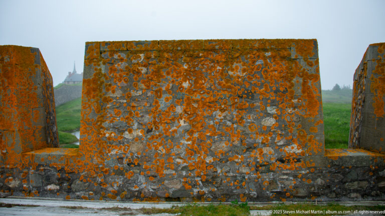 Lichen-covered stone parapet with broad rectangular openings, framing foggy grass and distant fortress buildings beyond.