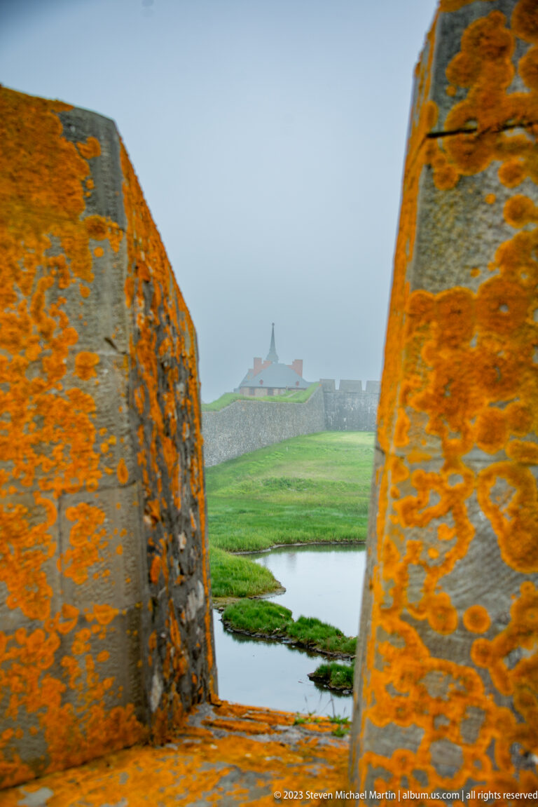 View through orange-lichen-covered stone walls toward a shallow ditch, green earthworks, and distant fortress buildings in mist.
