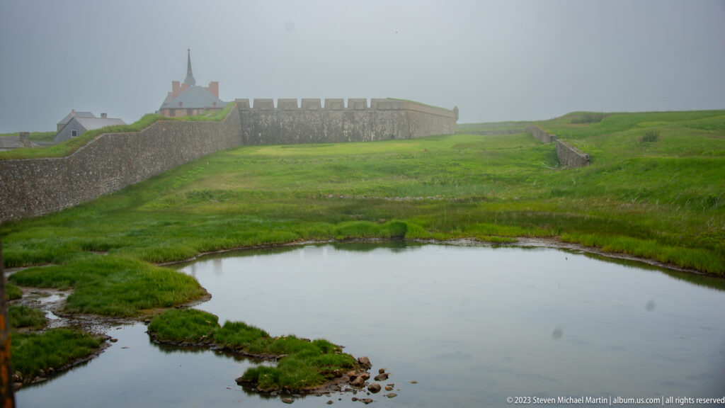 Water-filled defensive ditch bordered by green grass, with long stone fortress walls and misty buildings rising in the background.