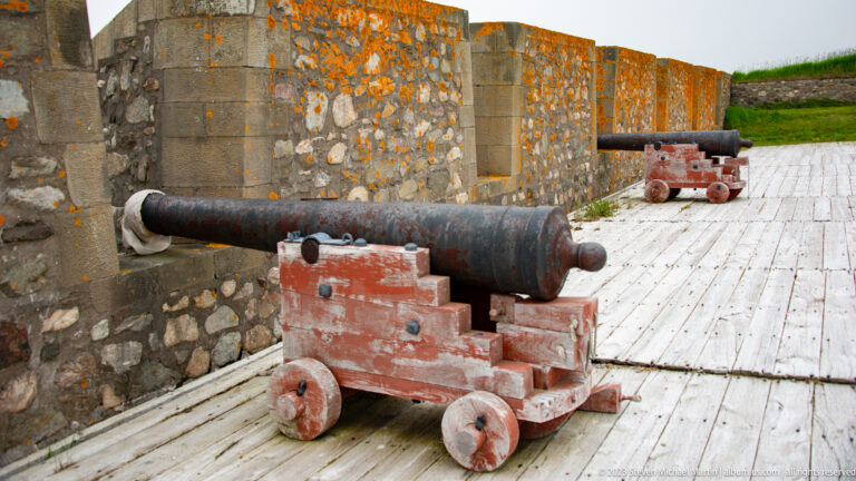 Black iron cannon on a red wooden carriage beside a lichen-covered stone wall, with a second cannon farther along the platform.
