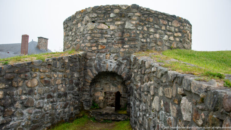 Circular stone structure set above a small arched passage within grassy fortress earthworks and rough masonry walls.