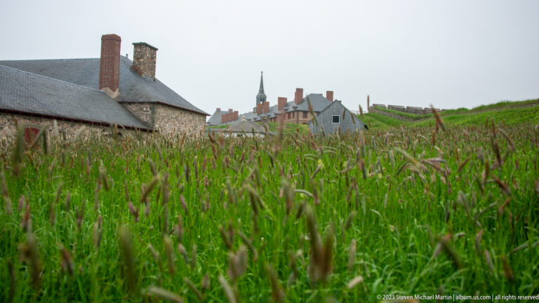 Tall green grass in the foreground with reconstructed stone buildings, chimneys, fencing, and a church spire beyond in mist.