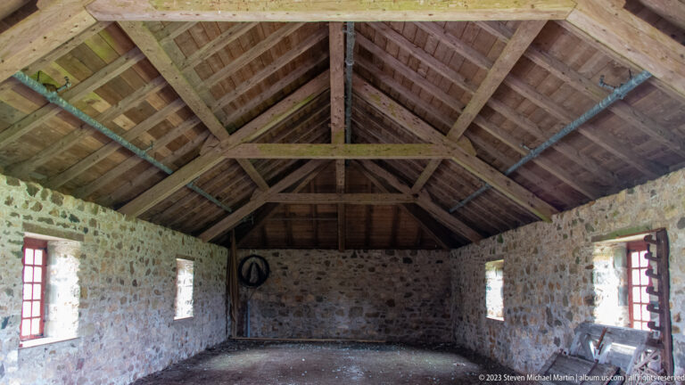 Empty stone room with red-trimmed windows and an exposed timber roof of rafters, purlins, and cross beams.