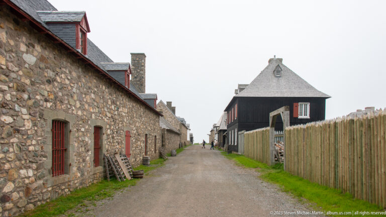Narrow gravel lane between a long stone building with red openings and a tall wooden palisade, with dark timber building ahead.