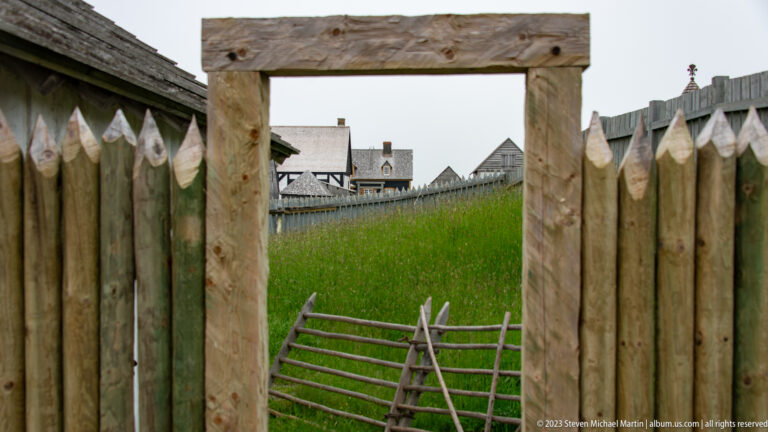 Rectangular opening in a pointed-log palisade framing a grassy slope, slatted gate, and reconstructed buildings beyond.