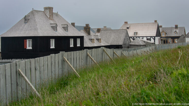 Black timber-clad building behind a gray wooden fence, with tall grass and a cluster of lighter historic buildings beyond.
