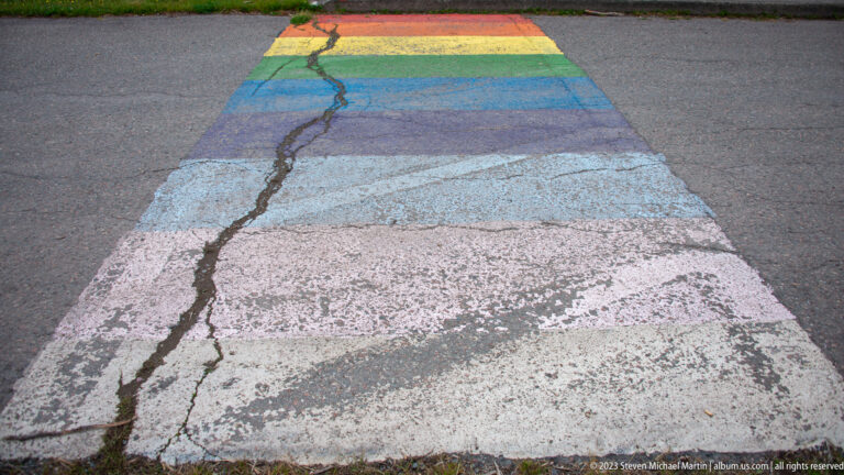 Weathered rainbow-painted pedestrian crossing extending across gray pavement.