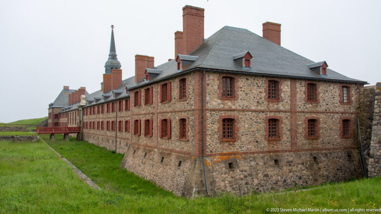 Large two-story stone building with red brick trim, many windows, steep gray roof, and tall chimneys beside the fortress town.