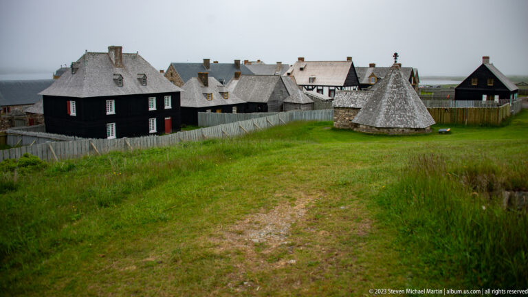 Reconstructed Louisbourg buildings, palisades, grassy earthworks, and a conical-roofed structure under an overcast coastal sky.