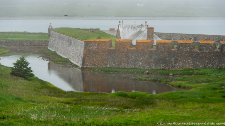 Stone fortress wall with turf-covered top beside a water-filled ditch and reconstructed buildings beyond.