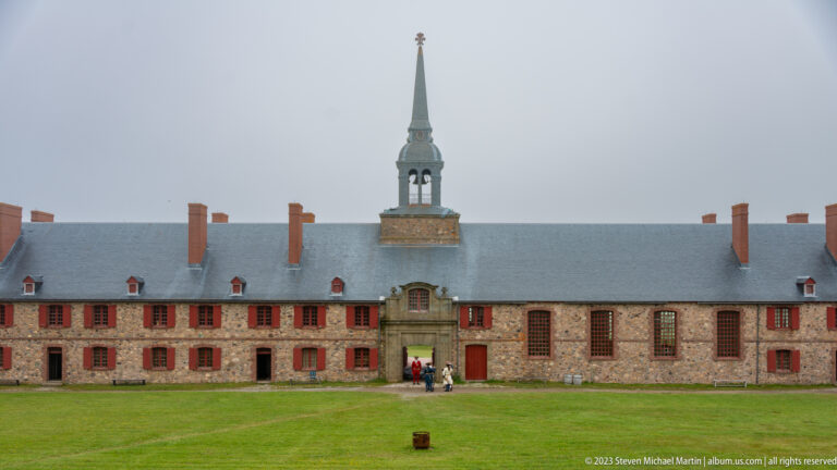 Long stone barracks with red shutters, slate roof, central doorway, and a tall chapel tower.