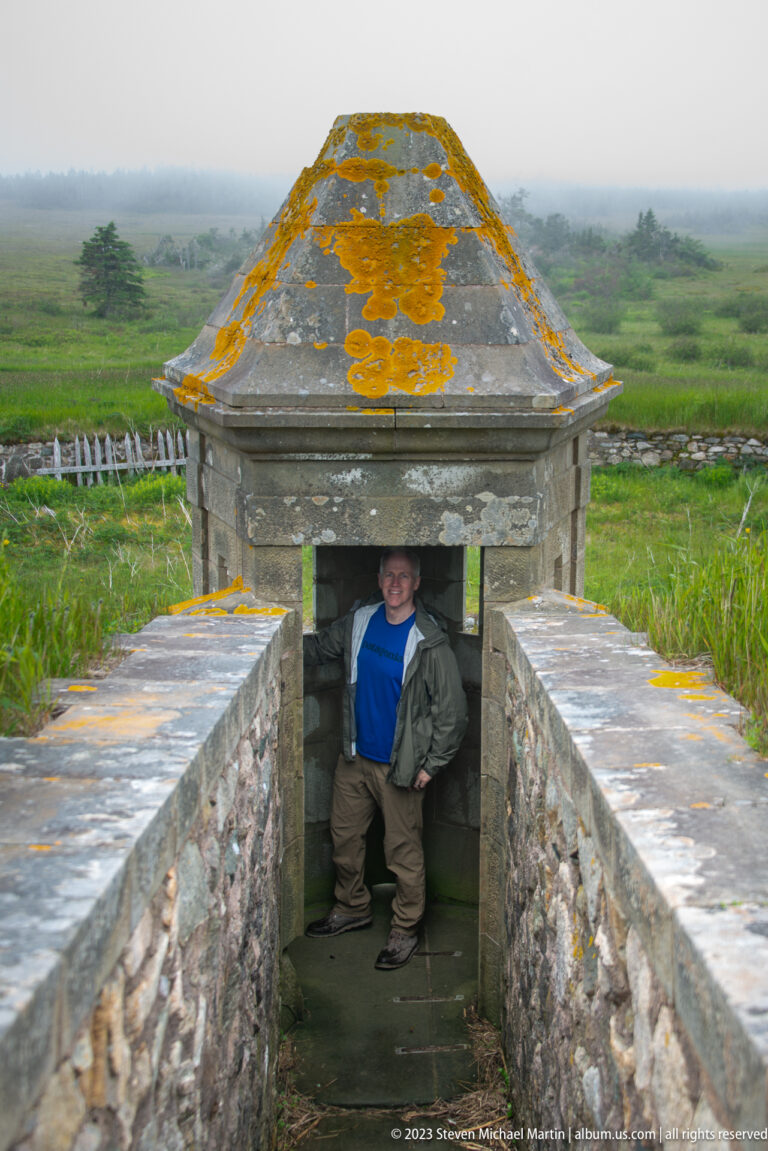 Small stone sentry box with steep lichen-covered roof and a seated person inside, framed by rampart walls.