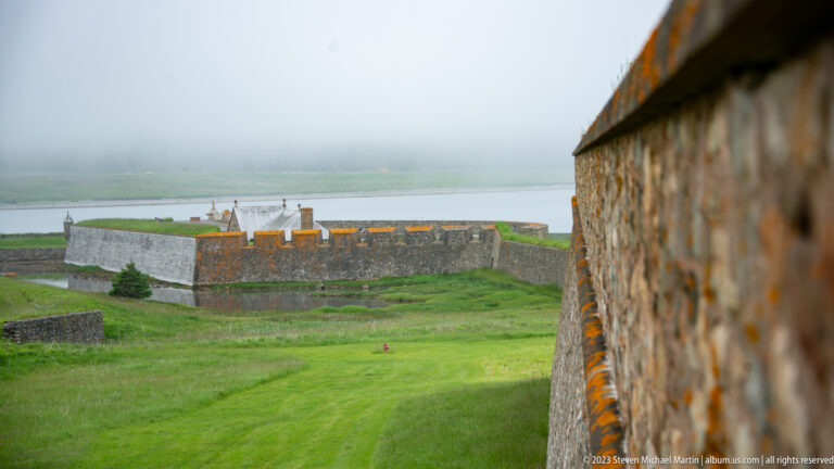 Fortress wall across a grassy defensive slope and ditch, viewed beside a close stone rampart at right.