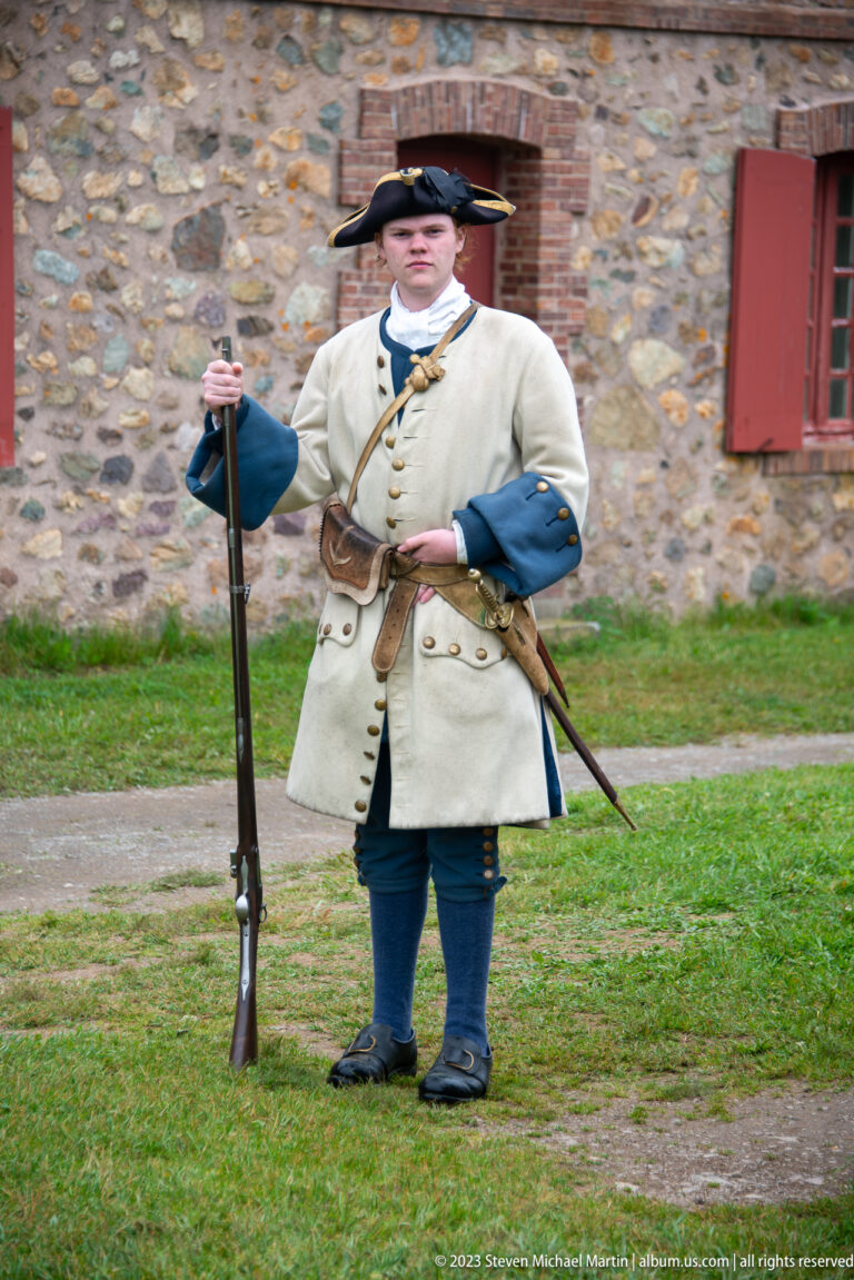 Costumed interpreter in tricorn hat and pale military coat holding a musket beside a stone building.