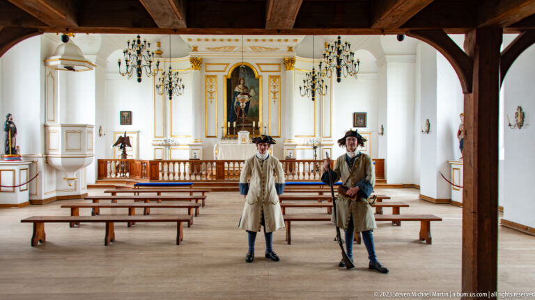 Two costumed interpreters standing among wooden benches inside a white chapel with gilded altar.