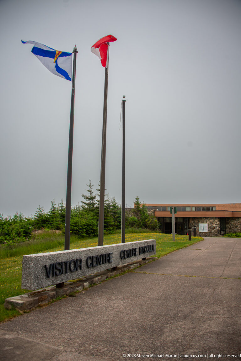 Visitor Centre approach with flagpoles, low bilingual stone sign, and modern building.
