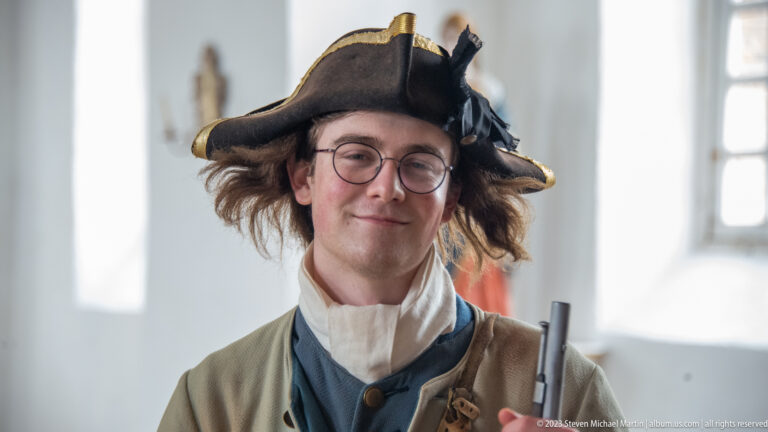 Close portrait of an interpreter with spectacles, tricorn hat, pale coat, blue waistcoat, and a musket at frame edge.