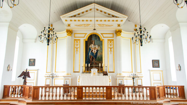 White and gold chapel altar with central religious painting, chandeliers, and wooden communion rail.