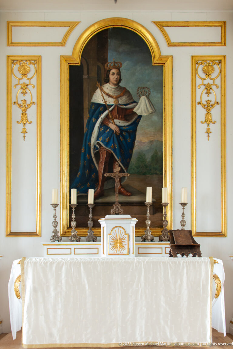 Large framed painting of a crowned figure above a white chapel altar with candles and gilded trim.