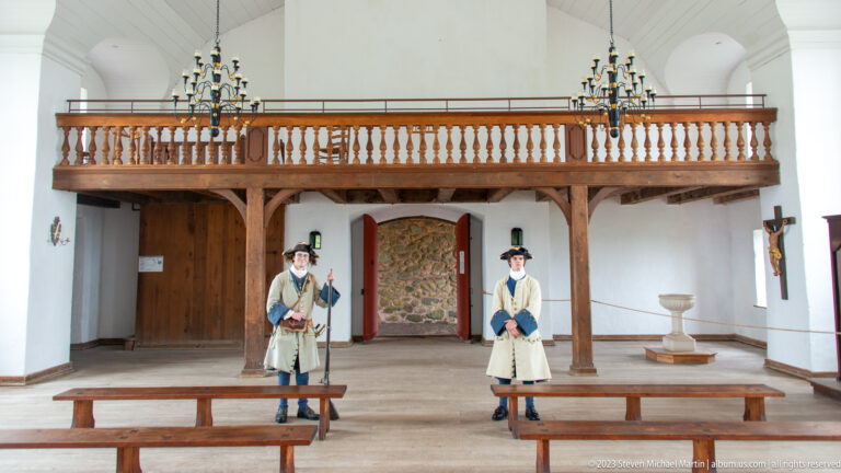 Two costumed interpreters beneath a wooden gallery inside the chapel, with benches and chandeliers.