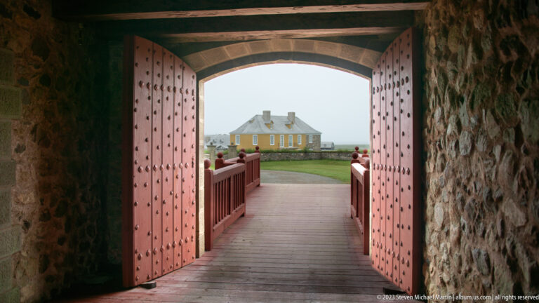 Open red studded fortress doors and timber passage framing a pale reconstructed building outside.