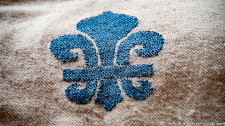 Close-up of a blue embroidered fleur-de-lis on coarse off-white woven cloth.