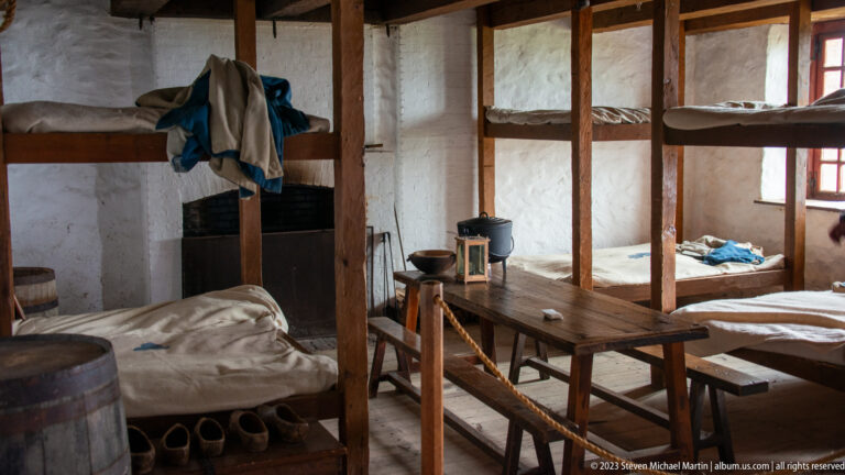 Wooden bunk beds, simple bedding, table, bench, barrel, and lantern inside a reconstructed soldiers’ barracks room.