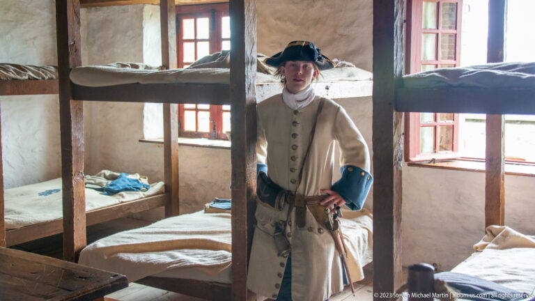 Costumed soldier in pale coat and tricorn hat standing among wooden bunk beds in a barracks room.