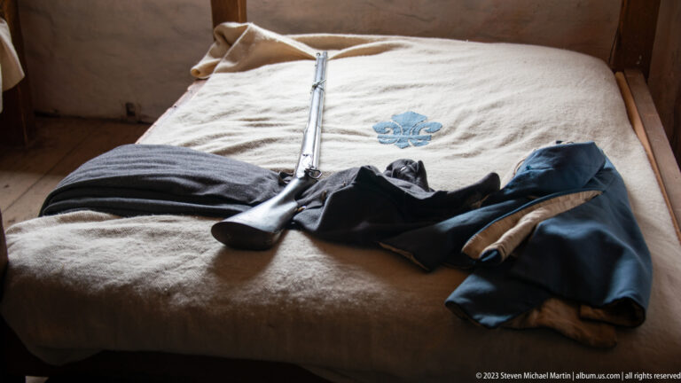 Musket, shoe, blue-and-pale uniform pieces, and bedding with blue fleur-de-lis arranged on a barracks bunk.