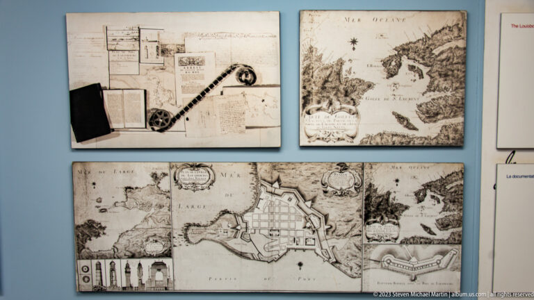 Interpretive display of historical maps, fortress plans, and coastal charts mounted on a pale blue wall.