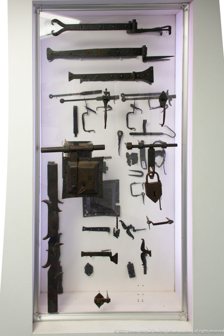 Backlit case containing corroded iron firearm parts, locks, tools, fasteners, and other metal artifacts.