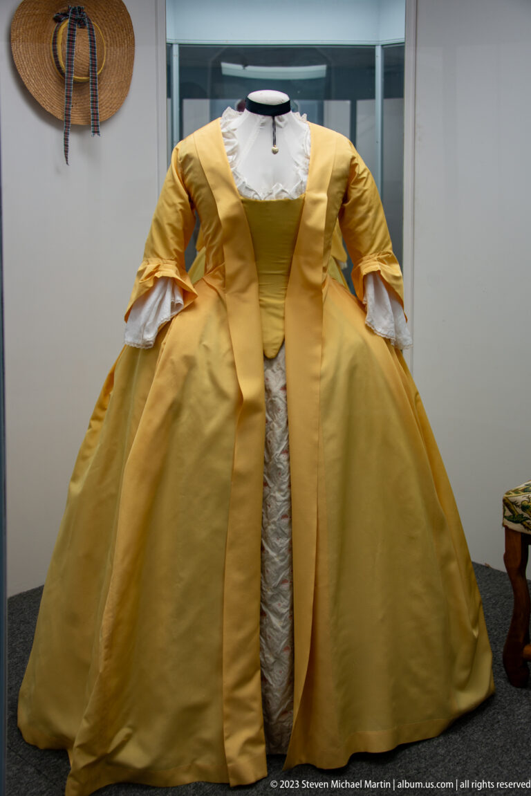 Yellow eighteenth-century-style women’s gown with wide skirt, fitted bodice, white cuffs, and pale central trim.