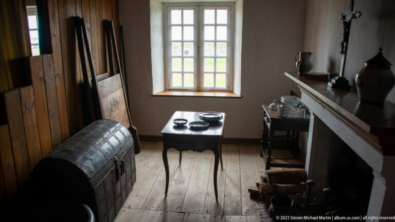 Wooden chest, small table with dishes, ceramic vessels, and hearth in a dim room lit by a multi-pane window.