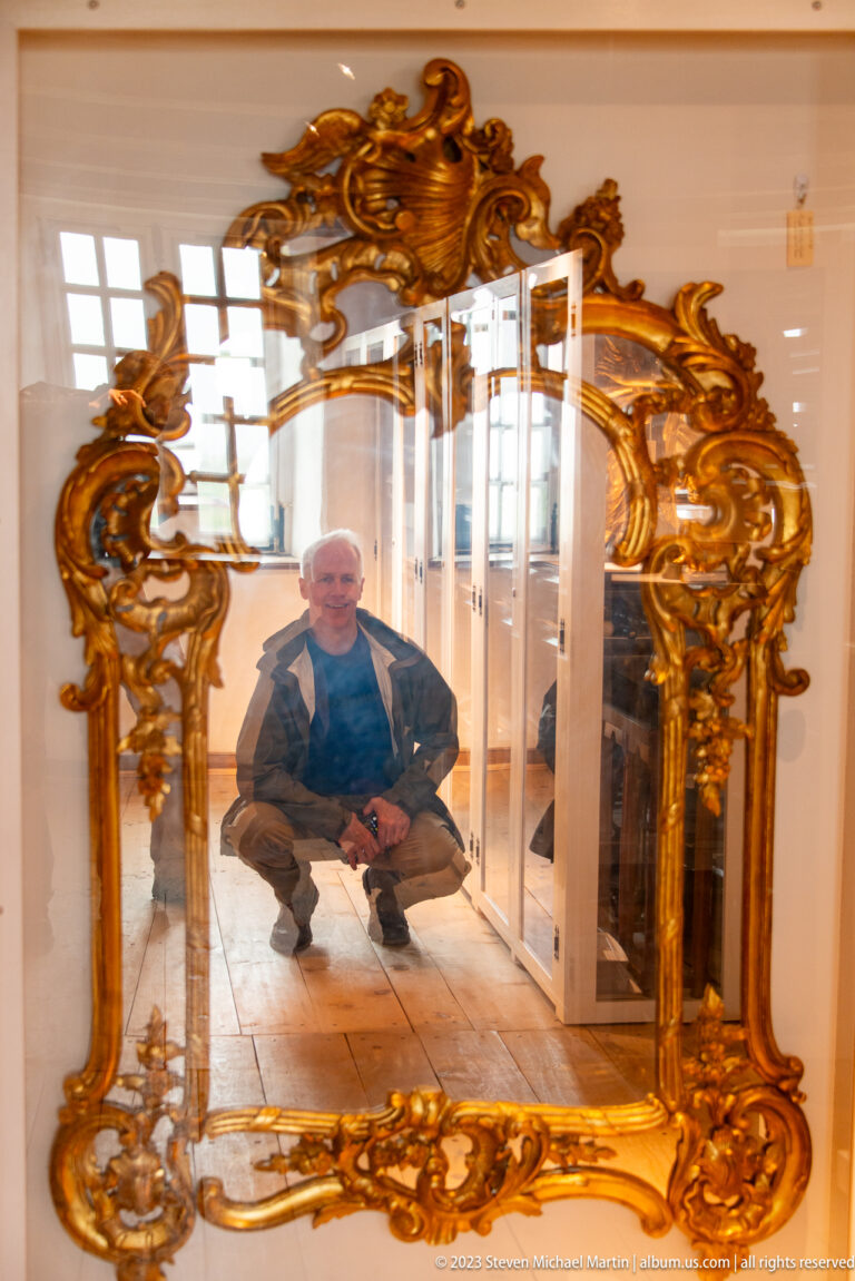 Large gilded rococo-style mirror with scrolling carved ornament, reflecting a seated visitor and bright museum interior.