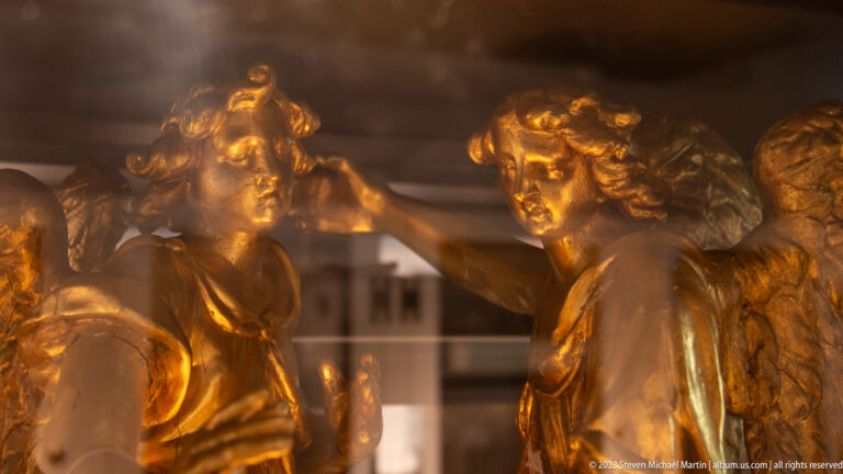 Close detail of two gilded winged figures facing each other, with modeled faces, curled hair, and feathered wings.