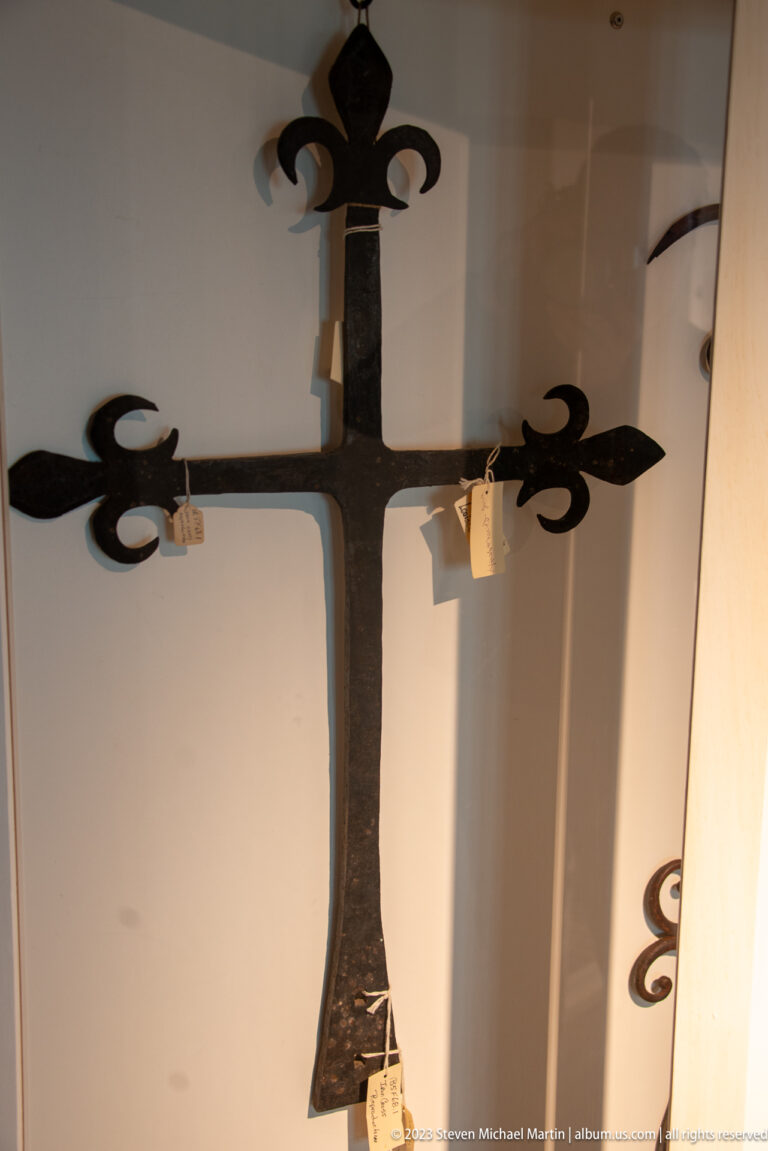 Forged iron cross with broad arms and fleur-de-lis terminals displayed against a pale museum wall.