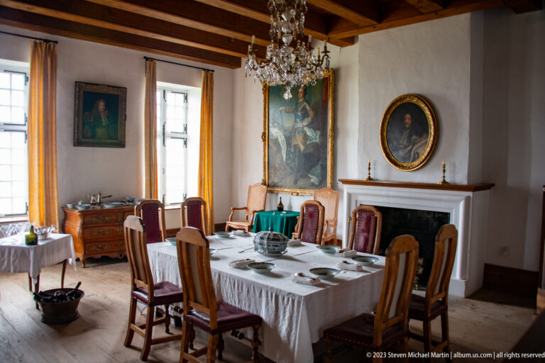 Reconstructed period dining room with set table, wooden chairs, chandelier, portraits, fireplace, and exposed ceiling beams.