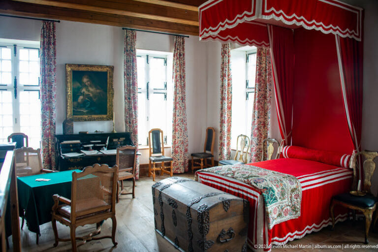 Reconstructed bedchamber with red canopy bed, patterned coverlet, large chest, writing table, chairs, and floral curtains.