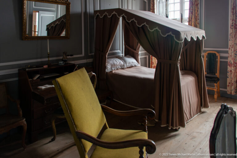 Curtained tester bed, upholstered armchair, desk, and floral window curtains in a reconstructed eighteenth-century bedchamber.