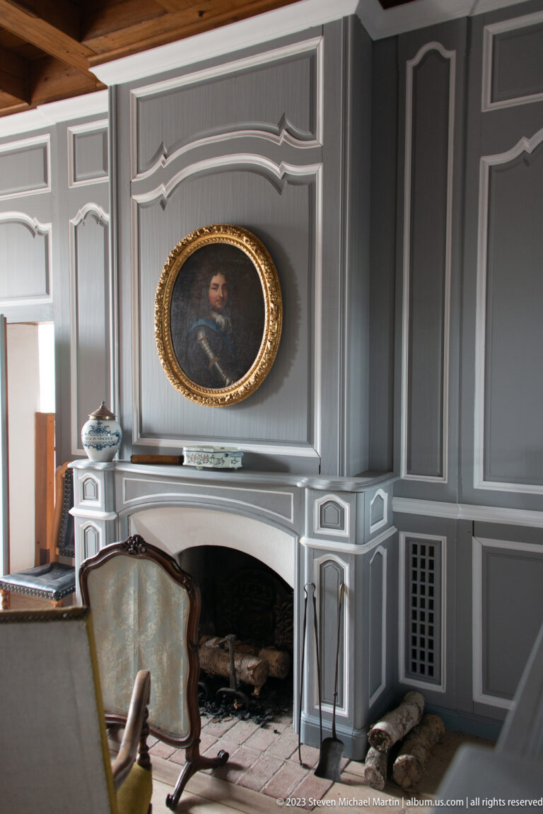 Gray paneled wall with white fireplace, gilded oval portrait, mantel objects, and upholstered chair in the foreground.