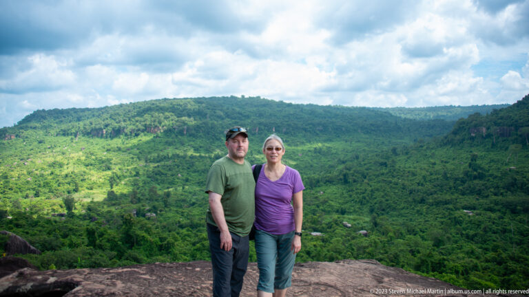 Two visitors stand on a rocky overlook above a broad green forested valley under clouds.