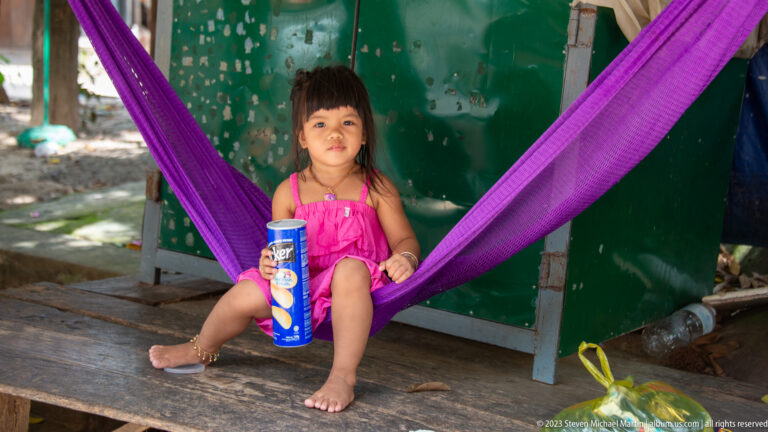 Young barefoot child in pink sits in a purple hammock holding a snack container.