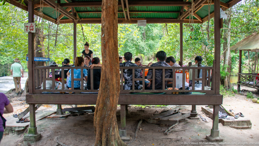 Visitors sit inside a raised open wooden pavilion surrounded by forest.