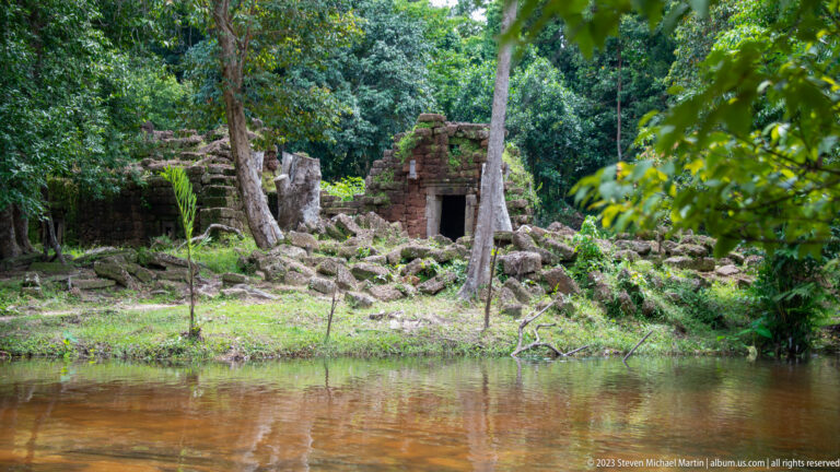 Ruined stone sanctuary with a dark doorway stands beyond a calm forest pool.