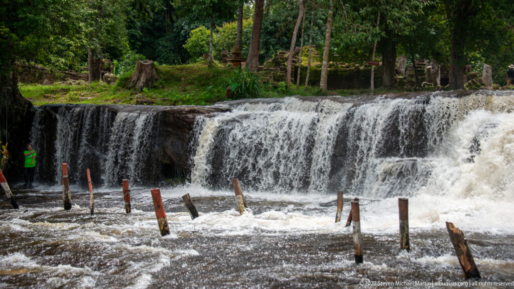 Broad low waterfall pours over a rock ledge with wooden posts in the foreground.