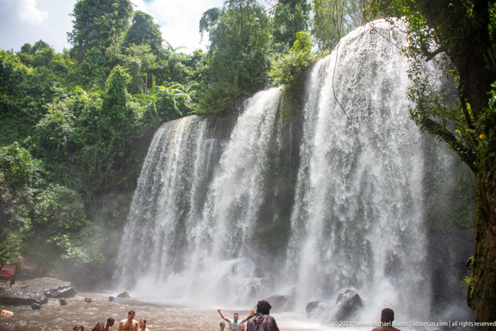Tall multi-curtain waterfall drops into a pool surrounded by dense tropical forest.