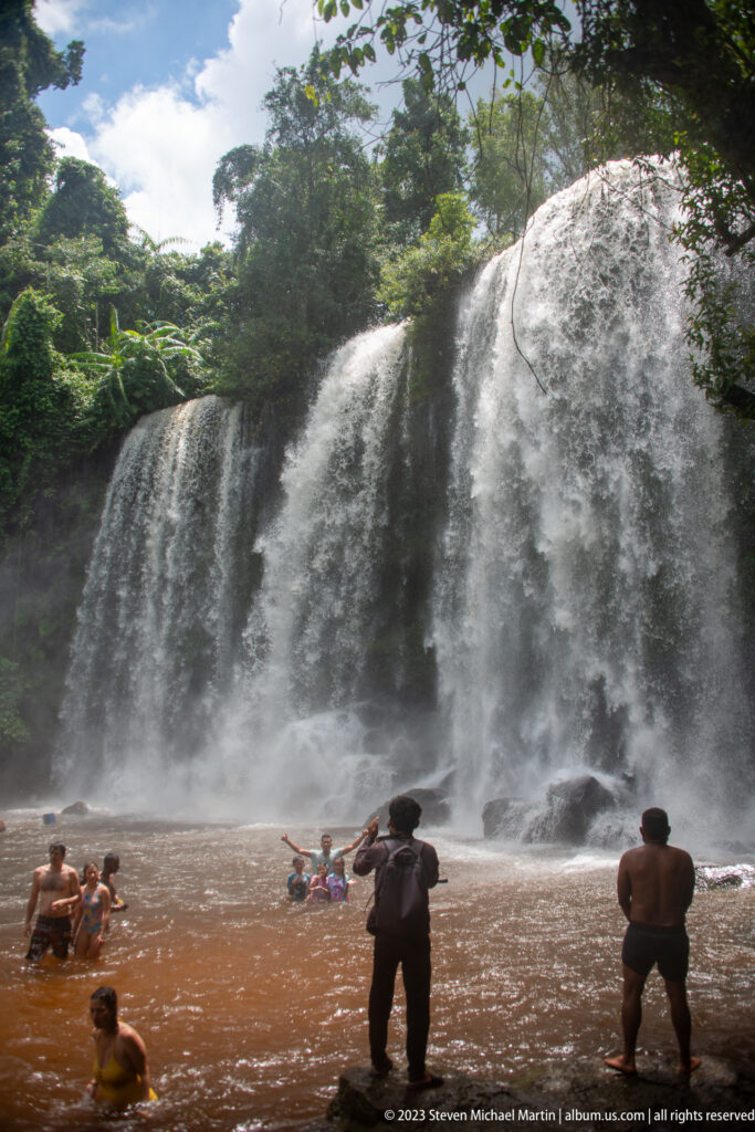 People swim and stand in a brown pool beneath a broad forest waterfall.