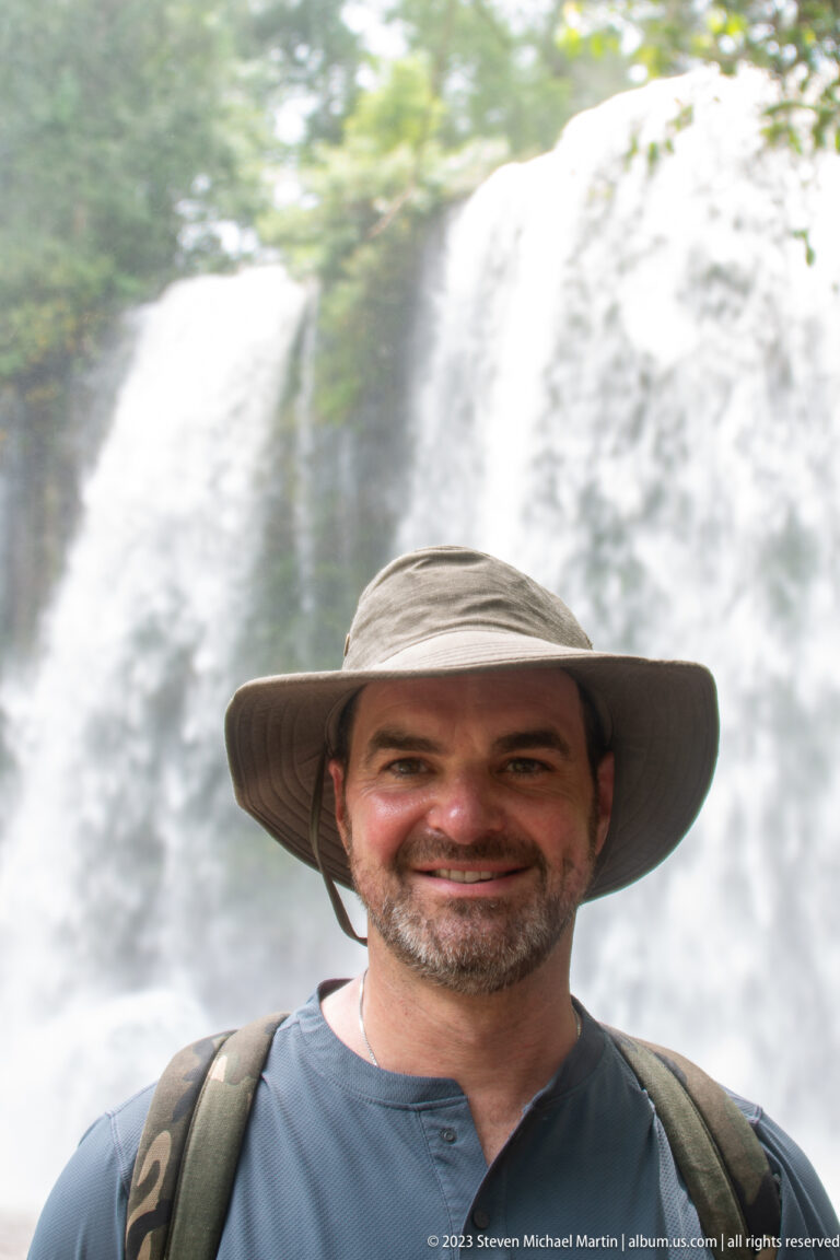 Bearded man in a wide-brimmed hat smiles with a waterfall blurred behind him.